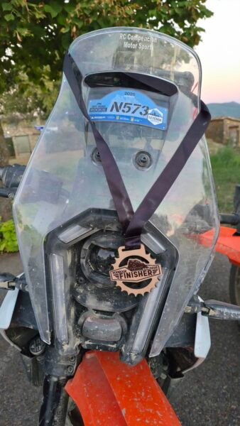 Medalla de madera Rodibook Finisher 2026 colgada del faro de una moto trail con el dorsal N573 en la cúpula
