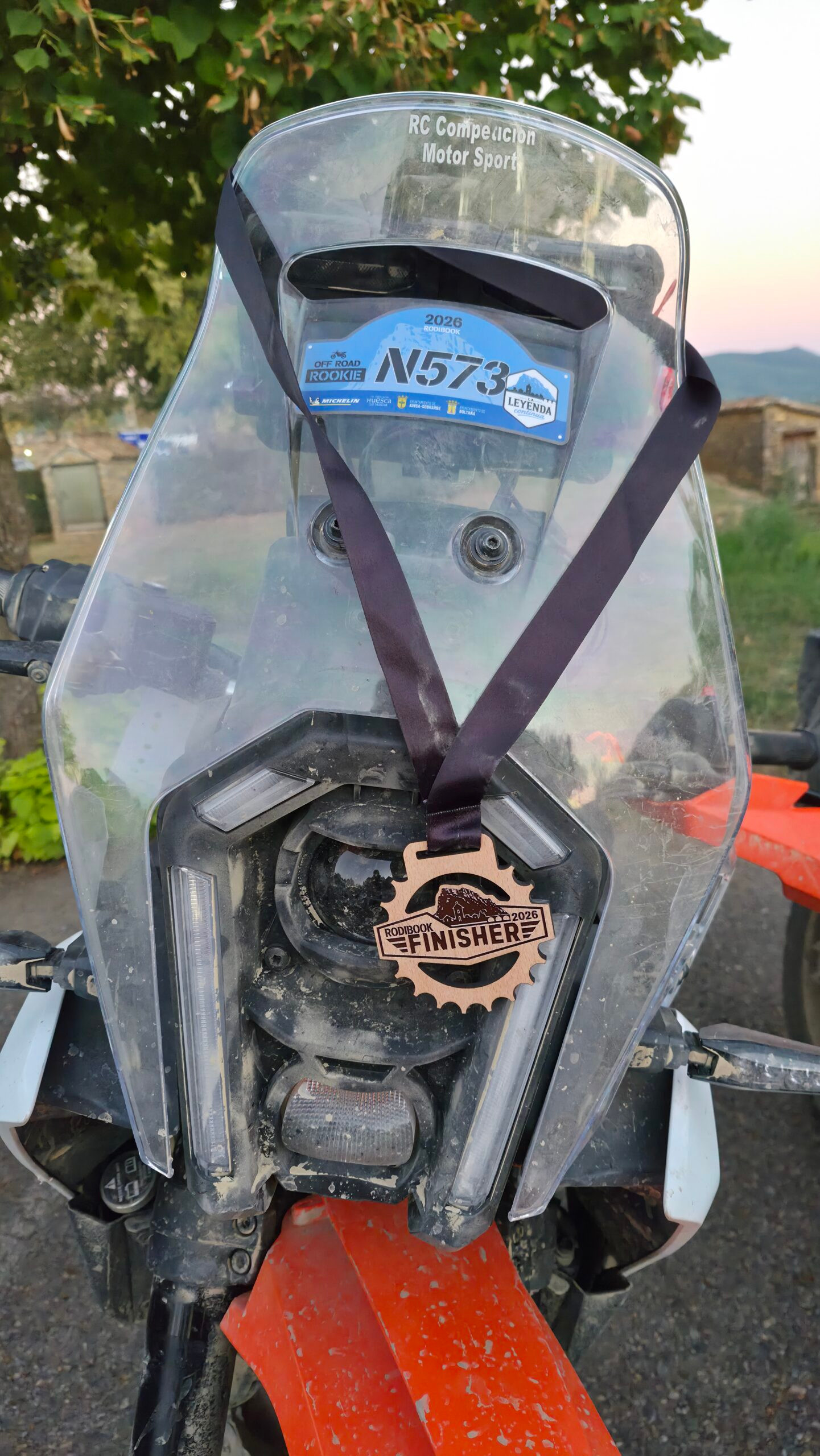 Medalla de madera Rodibook Finisher 2026 colgada del faro de una moto trail con el dorsal N573 en la cúpula